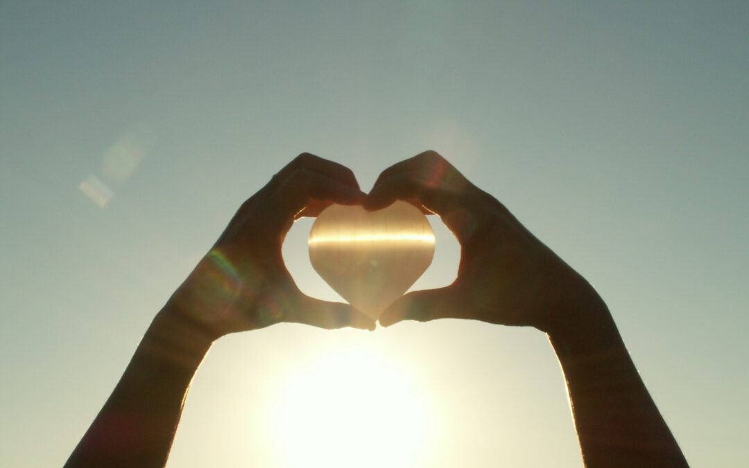 Two hands in heart shape holding a heart-shaped-stone in front of the sun.