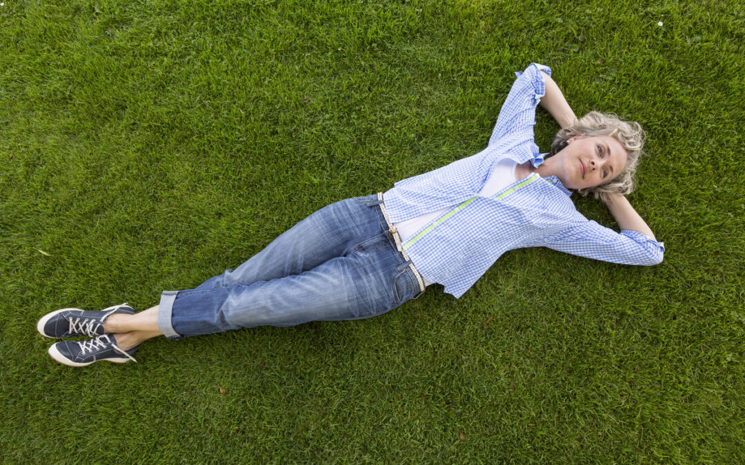 Middle-aged woman in casual weekend clothing relaxing on a grass lawn in a yard or park. She is smiling with a happy, contented expression and looks like she is daydreaming.