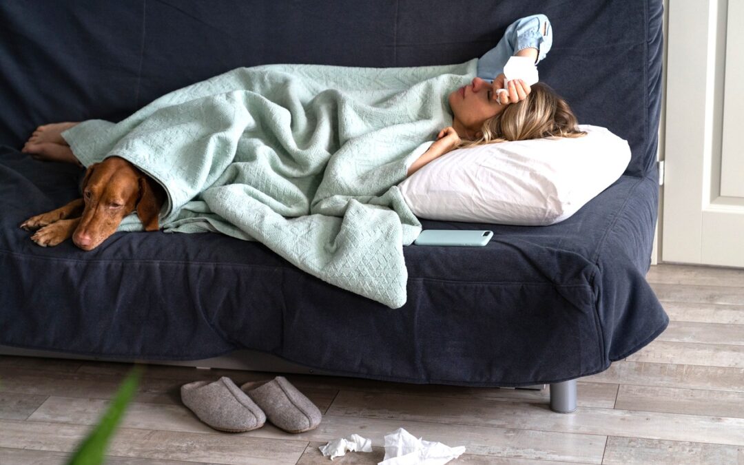 Sick woman on a couch under a blanket with her dog suffering from being sick,